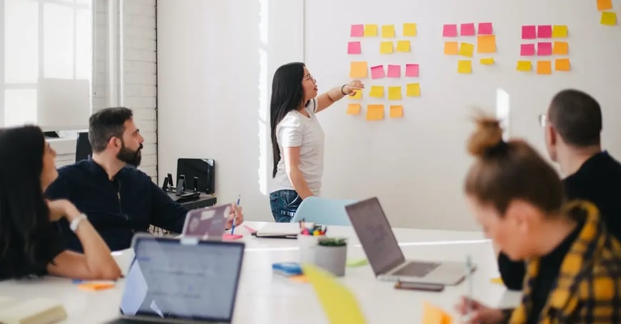 A team in a coference room discussing the in-person meeting budget posted on a whiteboard