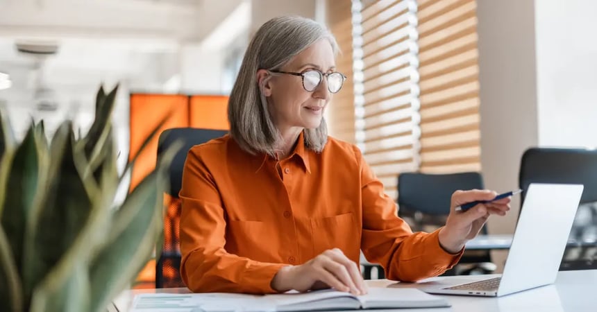 An older Executive Assistant sorting through meeting costs on her laptop wearing an organce shirt