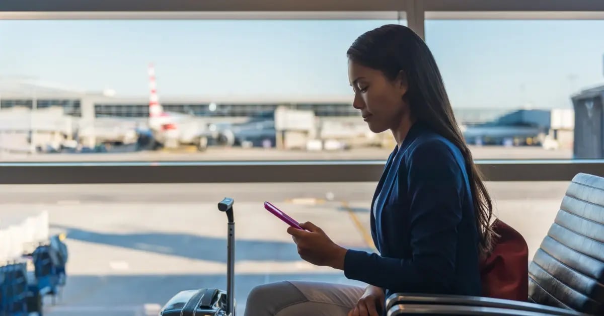 Attendee sitting in an airport chair with their suitcase and planes in the window behind them while on their phone