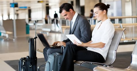Business travelers, a man and woman, sitting in airport seats with suitcases and working on their laptops