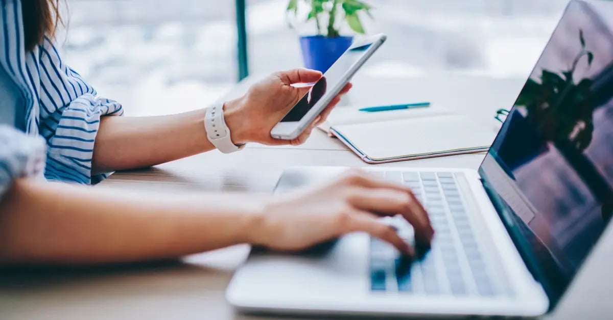 Cropped image of a woman holding a cell phone and typing on her laptop